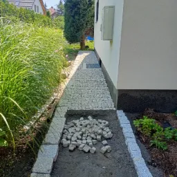 Ścieżka z szarej kostki granitowej w trakcie budowy obok białego budynku, widoczne ułożone obrzeża i stos kostki do dalszej pracy, wysoka trawa ozdobna w tle.