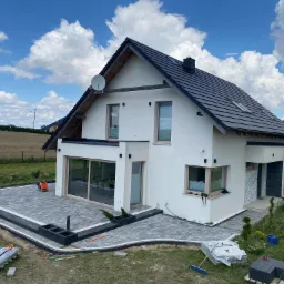 Nowoczesny dom z białymi ścianami i szarą kostką brukową na tarasie, widoczne narzędzia brukarskie i niezakończone prace, błękitne niebo z obłokami w tle.