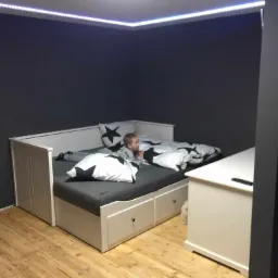 Chłopiec leży na białej rozkładanej sofie z szufladami na drewnianej podłodze, ściany pomalowane na ciemnoszaro, oświetlenie LED zamontowane pod sufitem.