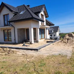 Nowoczesny dom jednorodzinny w trakcie budowy, widoczne przygotowane obrzeża chodnika z kostki w kolorze grafitowym, teren wokół w trakcie prac ziemnych, na drugim planie kolejne domy.