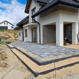 Tarasy z szarej kostki brukowej na podbudowie z piasku, widoczny pracownik przy maszynie i fragment domu w tle.