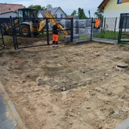 Przygotowanie terenu pod budowę w Skórczu: wyrównany teren piaskowy otoczony metalowym ogrodzeniem, w tle koparko-ładowarka JCB i palety z materiałami budowlanymi.