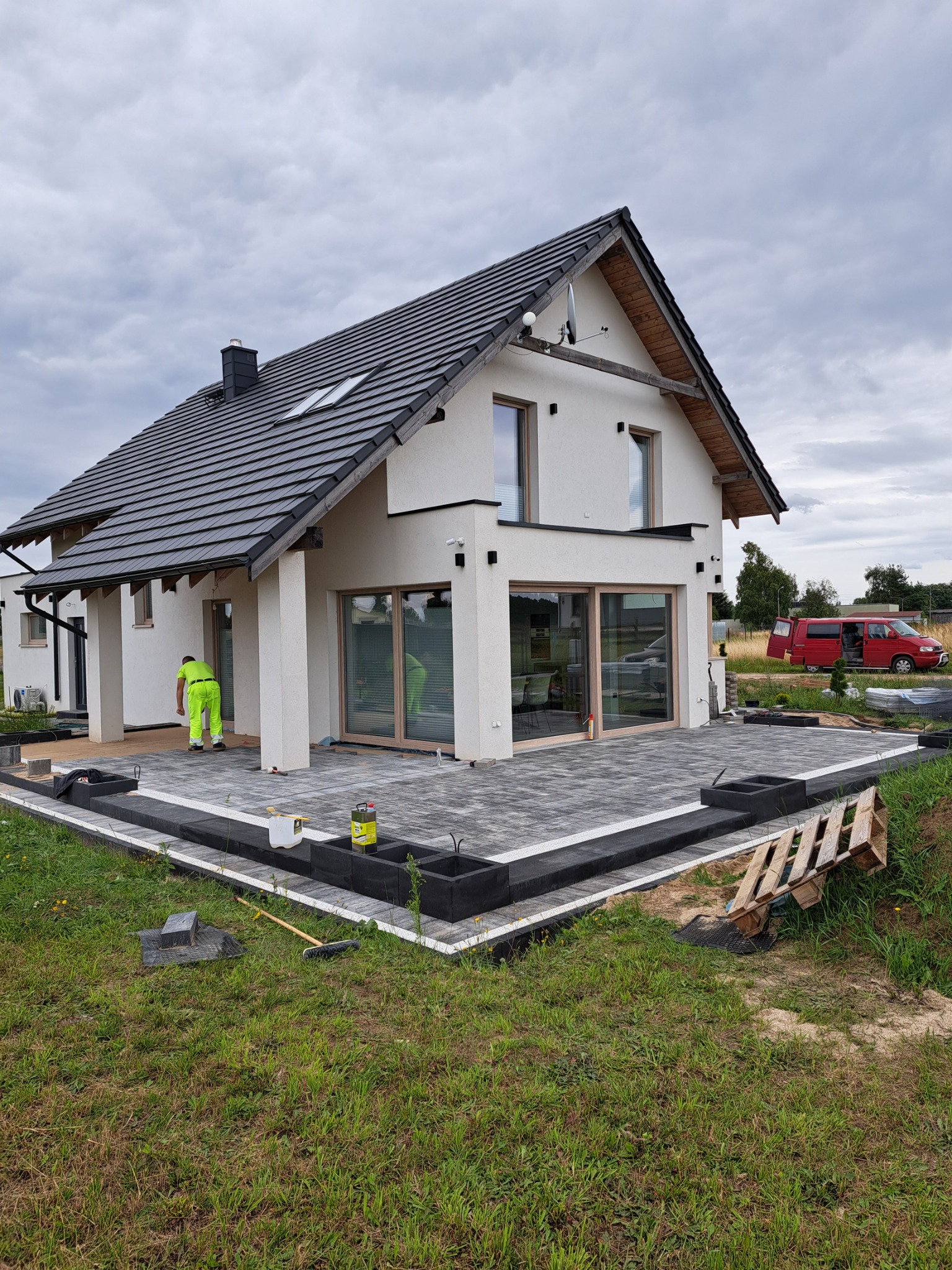 Nowoczesny dom z tarasem w trakcie budowy, widoczne ułożone szare kostki na tarasie, pracownik w jaskrawej odzieży roboczej oraz paleta i materiały budowlane w tle.