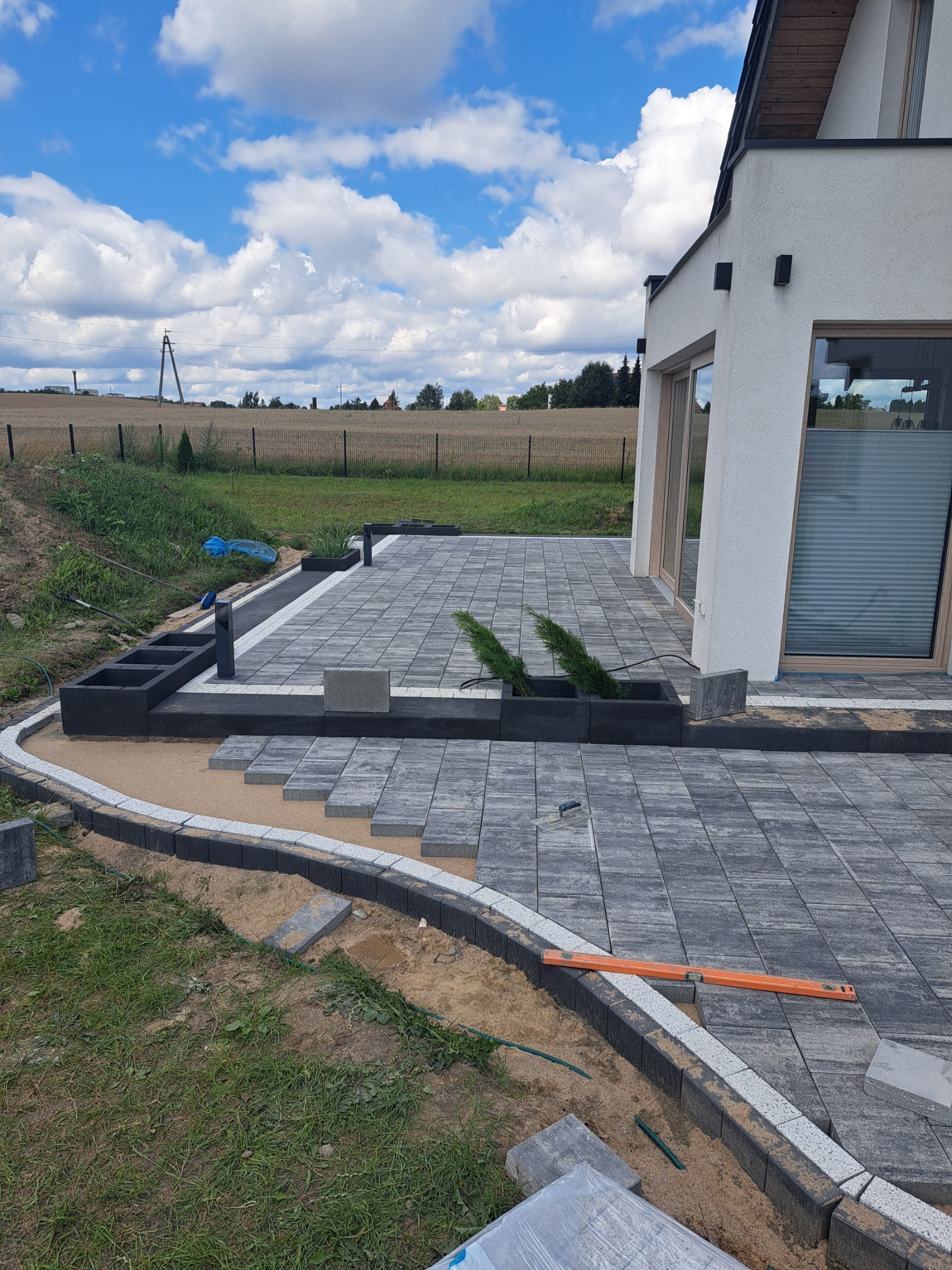 Fragment tarasu z szarej kostki brukowej w trakcie układania, widoczny poziomica, obrzeża z jasnych kamieni, w tle nowoczesny dom i pola uprawne.