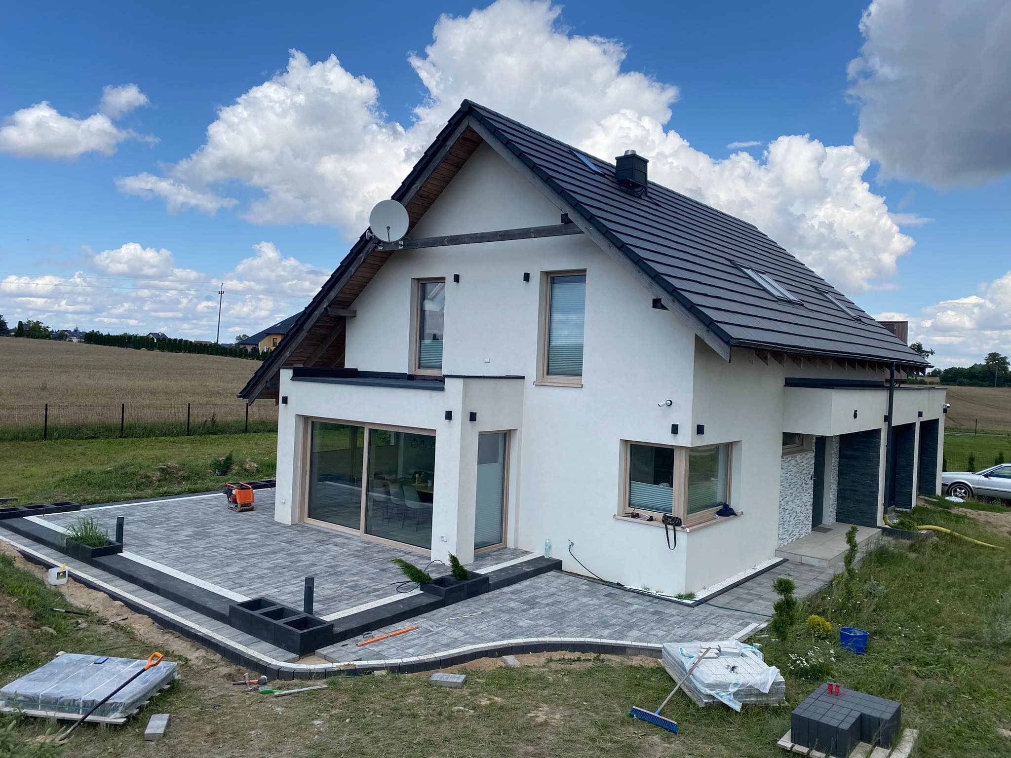 Nowoczesny dom z białymi ścianami i szarą kostką brukową na tarasie, widoczne narzędzia brukarskie i niezakończone prace, błękitne niebo z obłokami w tle.