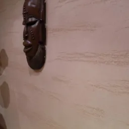 Teksturowana ściana w odcieniach beżu z dekoracyjną, drewnianą maską afrykańską, oświetlona ciepłym światłem