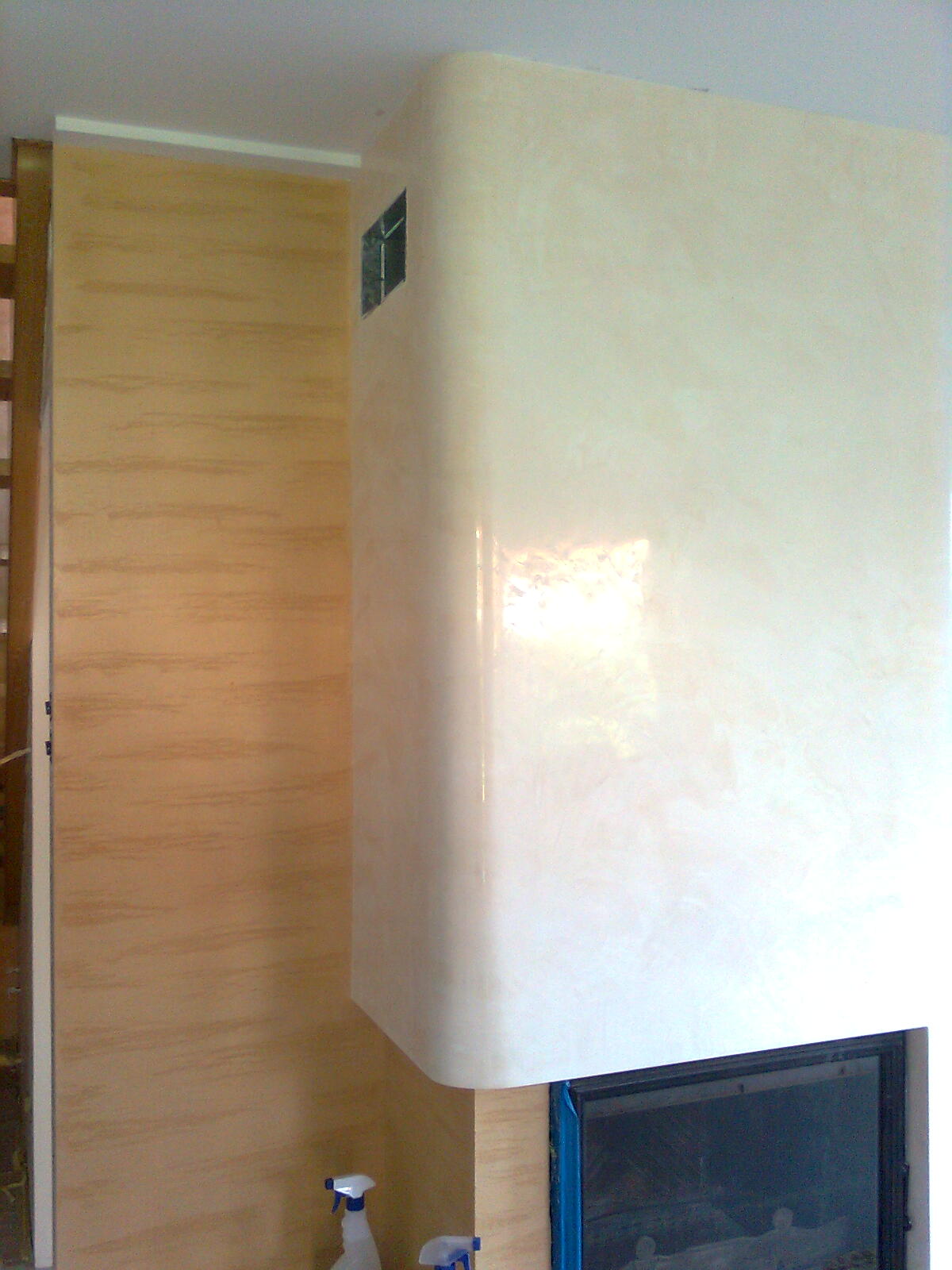 Biały, zaokrąglony kominek z wkładem, obok ściana z beżową tapetą strukturalną, widoczne butelki ze środkami czyszczącymi.