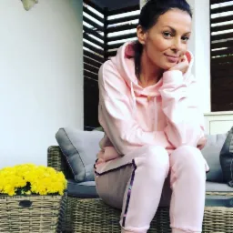 Kobieta siedząca na wiklinowym fotelu w różowym dresie z lampasami, obok bukiet żółtych chryzantem w wiklinowym koszu.