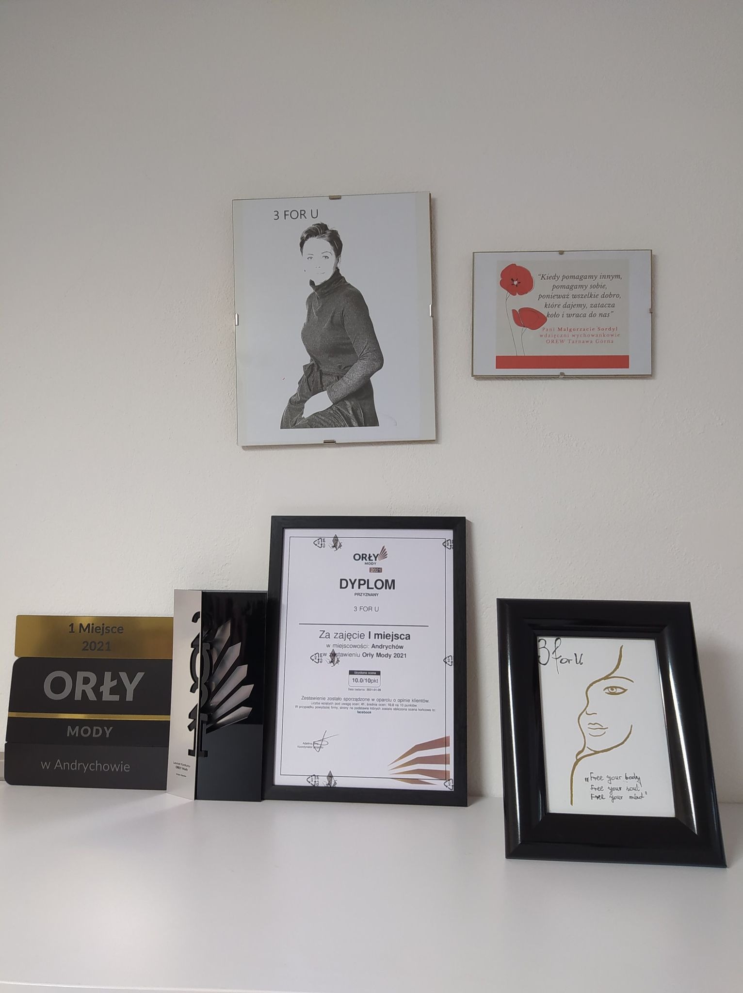 Nagrody i dyplom 'Orły Mody' firmy 3 FOR U na białej półce, w tle portret i sentencja w ramkach.