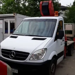 Biały samochód dostawczy Mercedes Sprinter z zamontowanym żurawiem dekarskim i pomarańczowym światłem ostrzegawczym, zaparkowany na placu budowy.