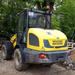 Żółto-szara ładowarka Wacker Neuson na żwirowym podłożu, z zielonymi drzewami w tle.