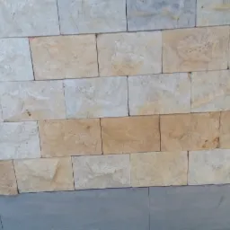 Ściana zewnętrzna wyłożona jasnymi, prostokątnymi płytkami imitującymi kamień, z widocznym fragmentem zieleni w tle.