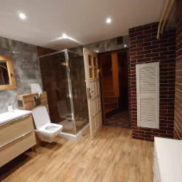 Rustykalna łazienka z drewnianą podłogą, kabiną prysznicową ze szklanymi drzwiami, toaletą podwieszaną, umywalką z szafką, lustrem w drewnianej ramie i ścianami wykończonymi płytkami imitującymi...