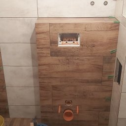 Grzegorz Marek - Montaż płytek imitujących drewno i jasnych płytek w łazience, widoczne krzyżyki dystansowe, otwór rewizyjny i rury kanalizacyjne.