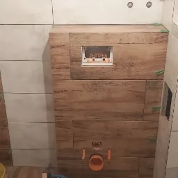 Montaż płytek imitujących drewno i jasnych płytek w łazience, widoczne krzyżyki dystansowe, otwór rewizyjny i rury kanalizacyjne.