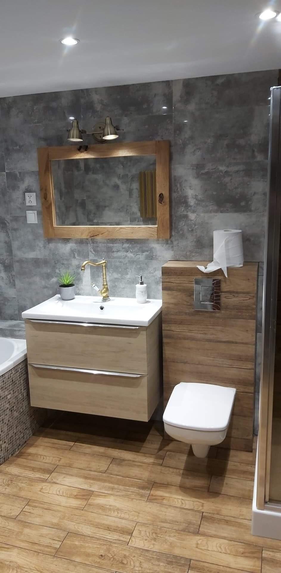 Nowoczesna łazienka z szarymi płytkami imitującymi beton, drewnianą podłogą, umywalką z szafką w jasnym drewnie, złotą baterią i lustrem w drewnianej ramie.