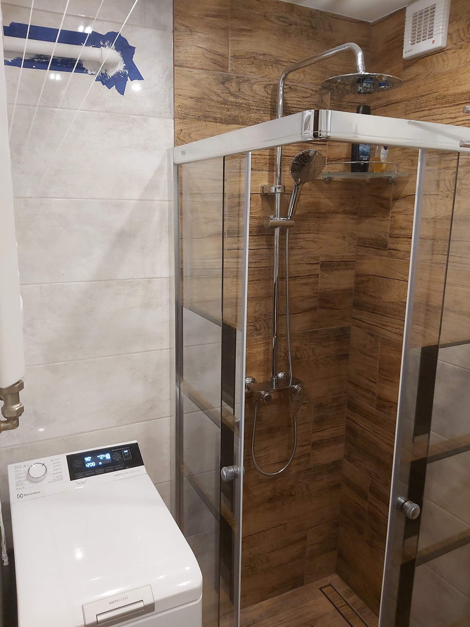 Nowoczesna kabina prysznicowa z deszczownicą i półką na kosmetyki, obok pralka Electrolux, drewnopodobne płytki na ścianach, widoczne ślady po montażu karnisza zabezpieczone taśmą.