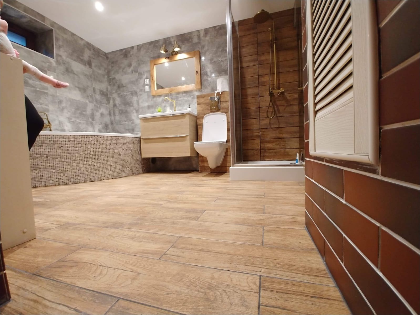 Nowoczesna łazienka z drewnopodobnymi płytkami na podłodze i ścianach kabiny prysznicowej, szare płytki na ścianie z wanną, wisząca szafka z umywalką i lustrem z oświetleniem, toaleta typu Geberit,...