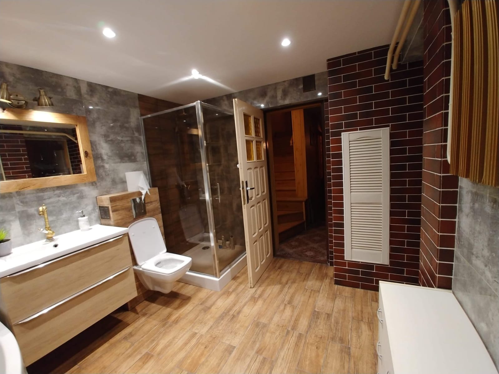 Rustykalna łazienka z drewnianą podłogą, kabiną prysznicową ze szklanymi drzwiami, toaletą podwieszaną, umywalką z szafką, lustrem w drewnianej ramie i ścianami wykończonymi płytkami imitującymi...