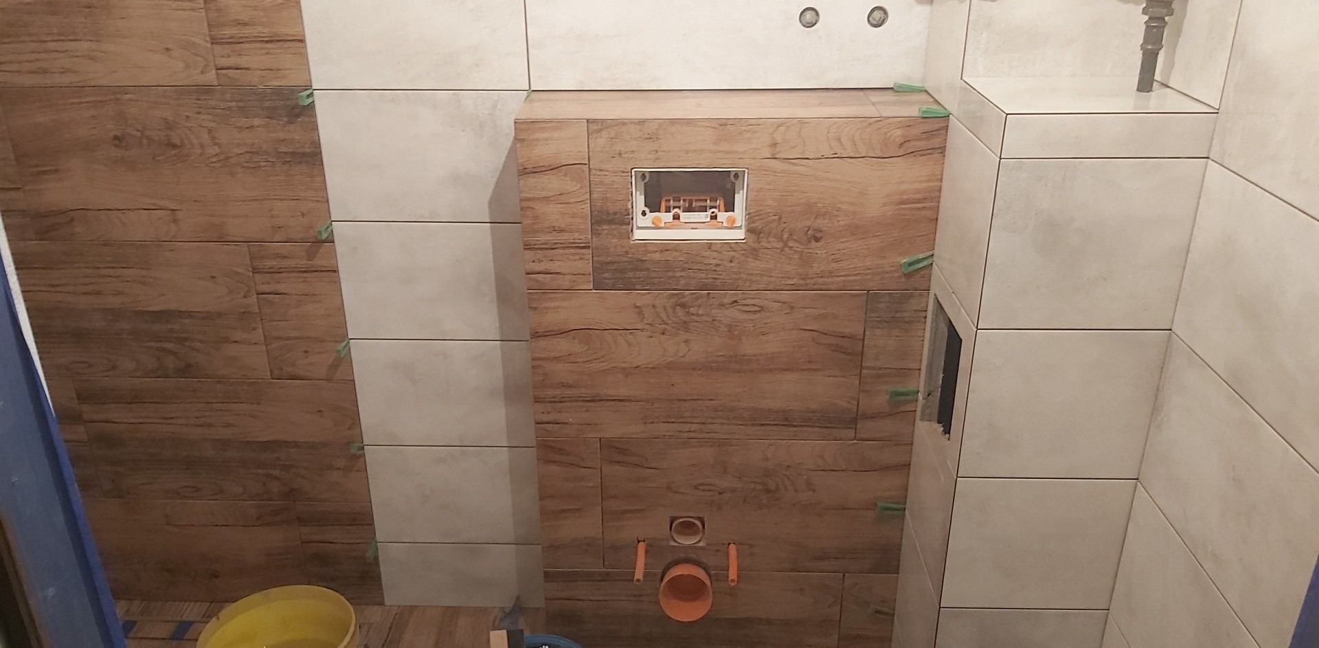 Montaż płytek imitujących drewno i jasnych płytek w łazience, widoczne krzyżyki dystansowe, otwór rewizyjny i rury kanalizacyjne.