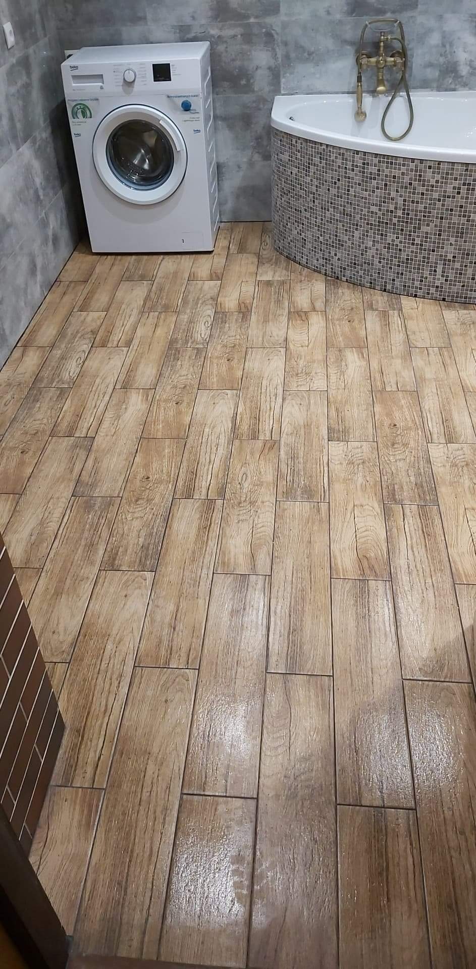 Ukończona łazienka z drewnopodobną podłogą, mozaikową obudową wanny i pralką Beko.