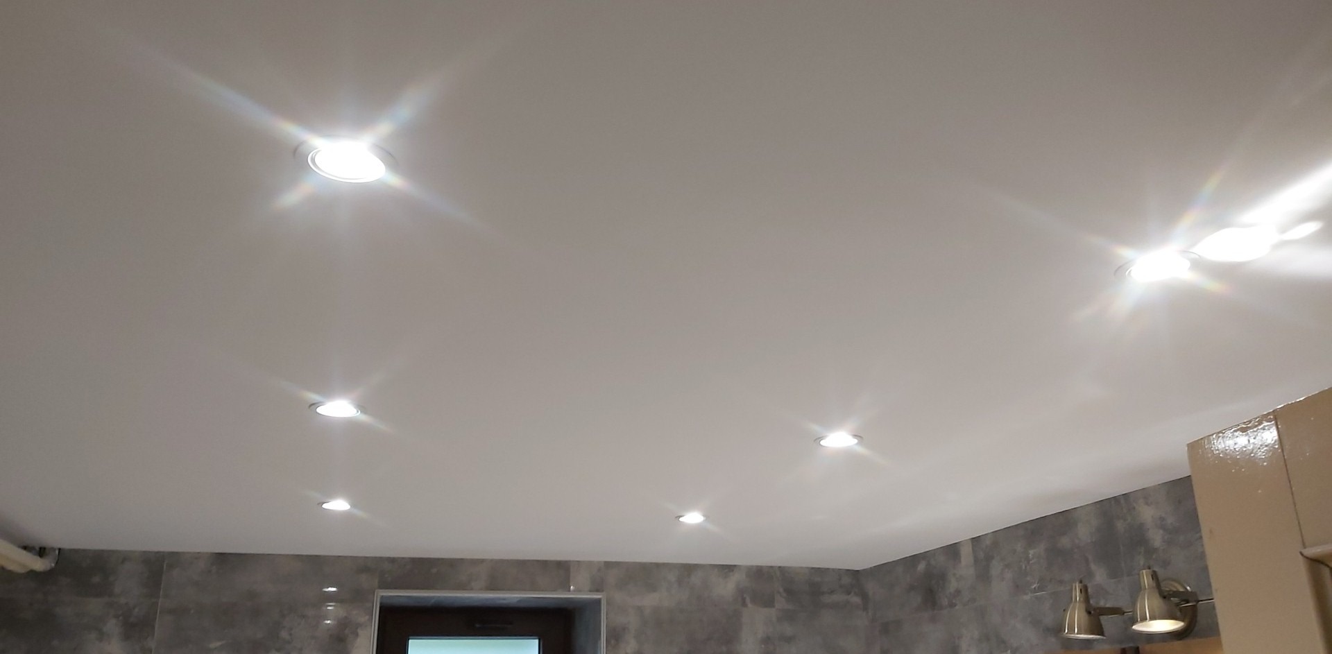 Biały sufit napinany z sześcioma punktowymi lampami halogenowymi, szare płytki na ścianie, fragment szafki i podwójny kinkiet w łazience.