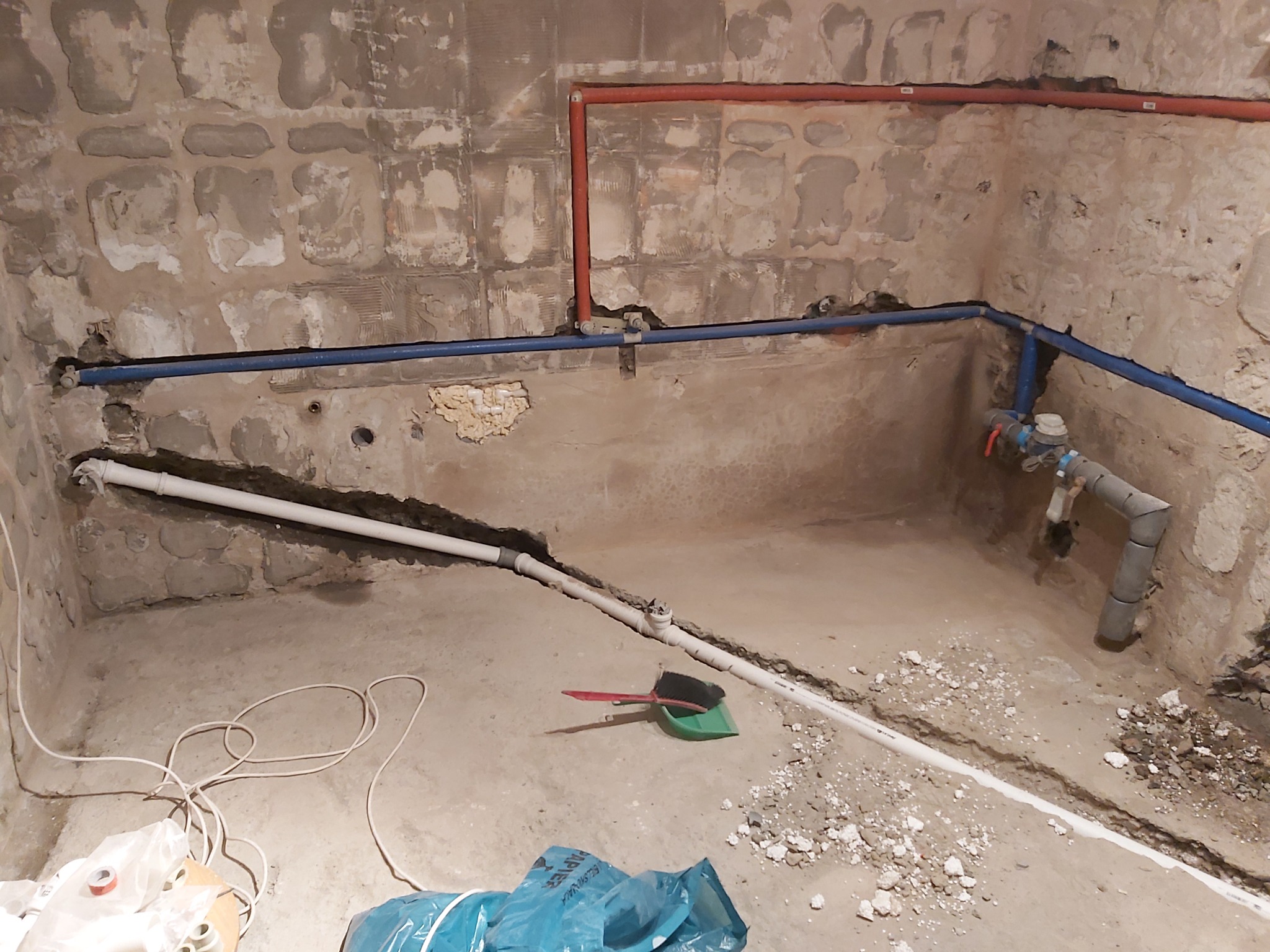 Surowa łazienka w trakcie prac instalacyjnych: widoczne odkryte rury wodne w kolorze czerwonym i niebieskim, białe rury kanalizacyjne w bruzdach w podłodze i ścianach, oraz narzędzia do sprzątania.