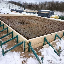 Zalany śniegiem szalunek pod fundament z siatką zbrojeniową, widoczne zielone podpory i rury kanalizacyjne wewnątrz konstrukcji.