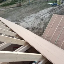 Konstrukcja drewniana dachu w trakcie budowy, widoczne elementy szkieletu i płyty poszycia zewnętrznego z brązową membraną w tle.