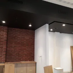 Ciemny sufit podwieszany z wbudowanymi punktowymi lampami w pomieszczeniu z fragmentem ściany z czerwonej cegły i białymi ścianami, widoczny pracownik w tle.