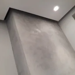 Ściana wykończona dekoracyjnym tynkiem w odcieniach szarości, z widocznym oświetleniem punktowym i fragmentem sufitu z czarną listwą wykończeniową.