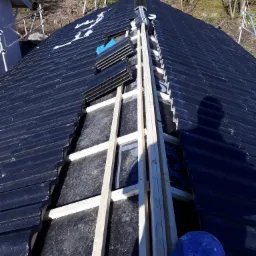 Montaż czarnej dachówki ceramicznej na drewnianej konstrukcji dachu, widok z góry, z boku leży niebieska czapka.