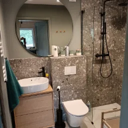 Nowoczesna łazienka w Wejherowie z okrągłym lustrem, umywalką nablatową na drewnianej szafce, toaletą i kabiną prysznicową z czarną armaturą na tle ściany wykończonej płytkami imitującymi lastryko.