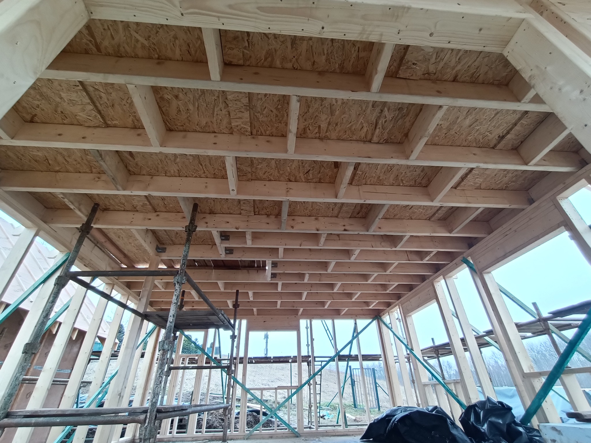 Wnętrze domu w trakcie budowy w technologii szkieletowej, widok na drewnianą konstrukcję stropu i ściany z widocznymi elementami rusztowania.