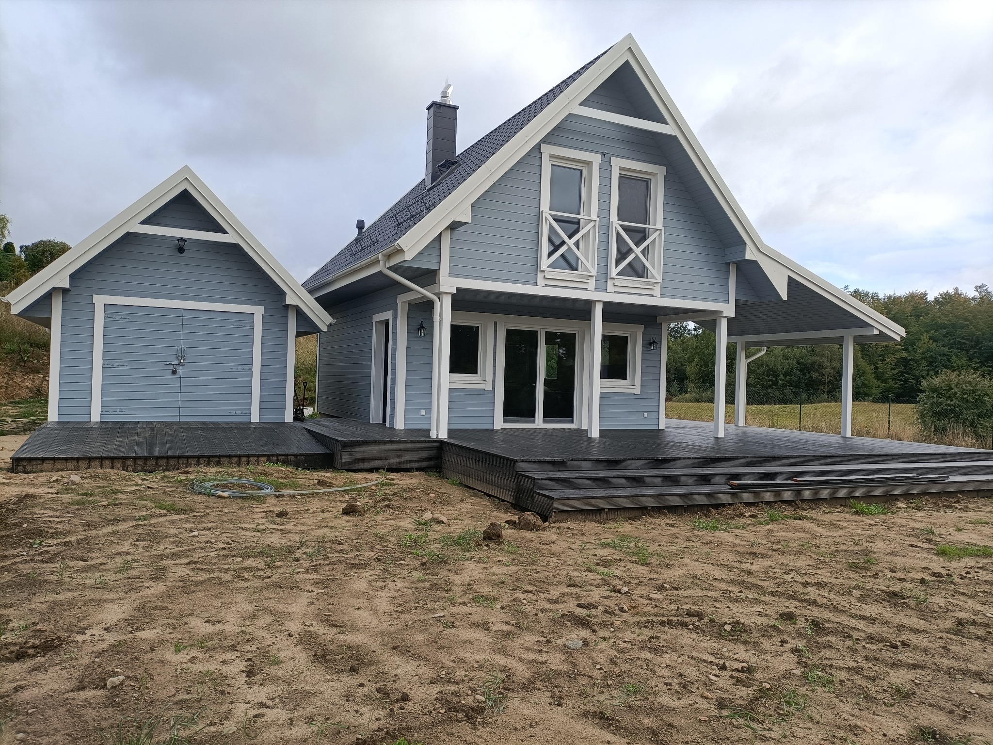 Niebieski dom szkieletowy z ciemnym dachem i drewnianym tarasem, widok z przodu na tle pochmurnego nieba i niezagospodarowanego terenu.