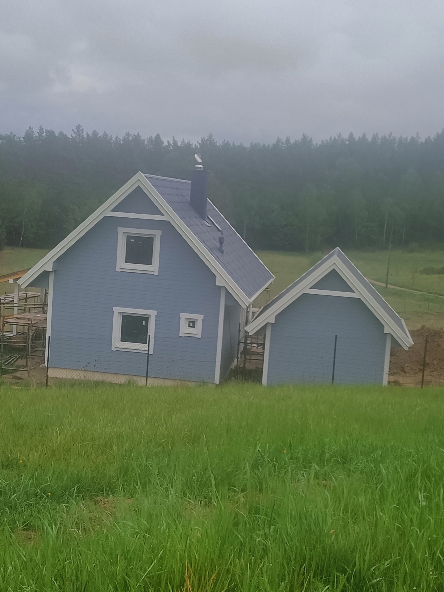 Niebieski dom z szarym dachem w trakcie budowy na tle zalesionego wzgórza, widoczne rusztowania i świeżo posiana trawa.