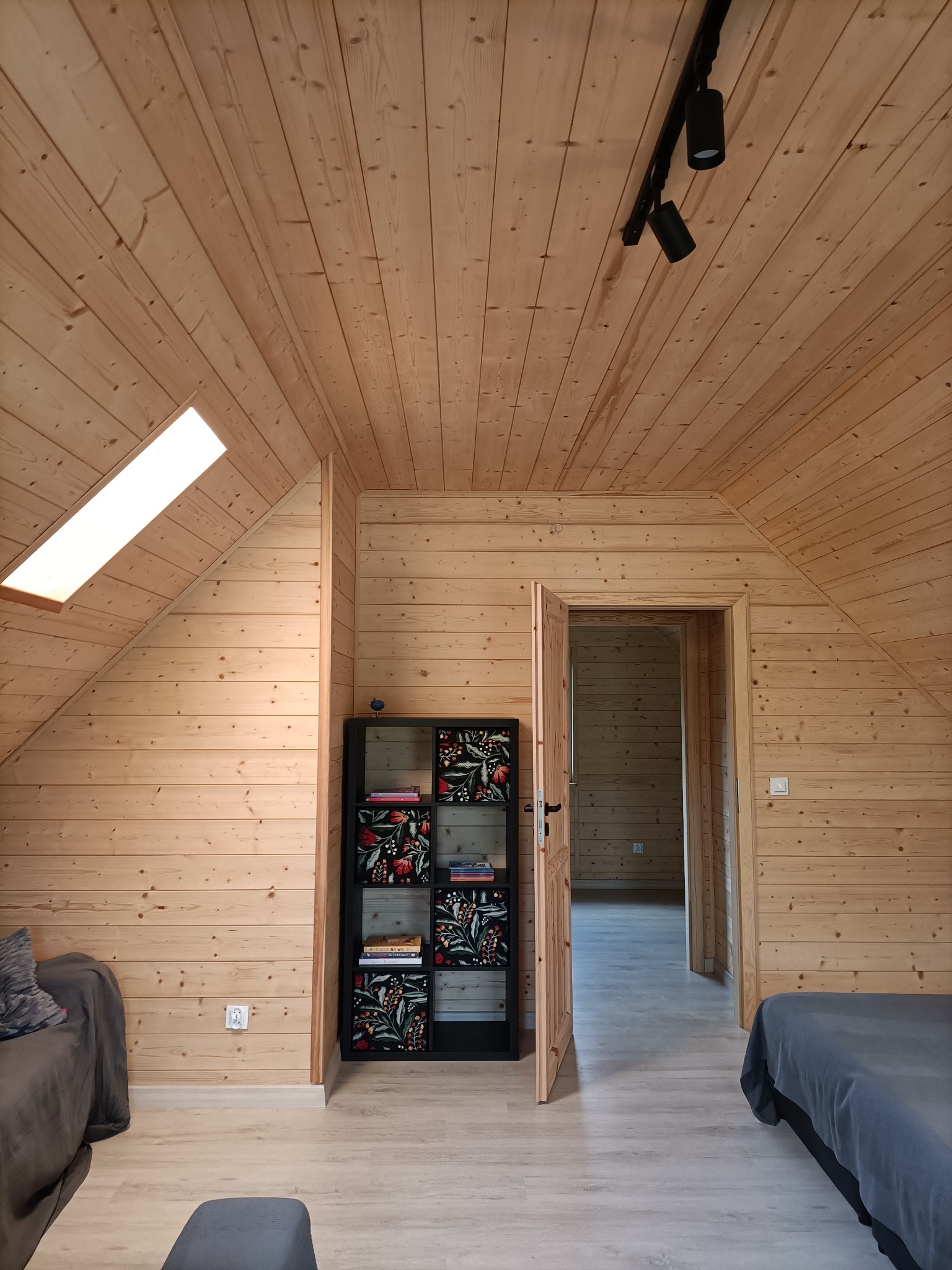 Wnętrze poddasza wykończonego drewnem, widoczne drewniane ściany, sufit z oknem dachowym, oświetlenie szynowe, regał z wzorzystymi panelami, częściowo widoczne meble.