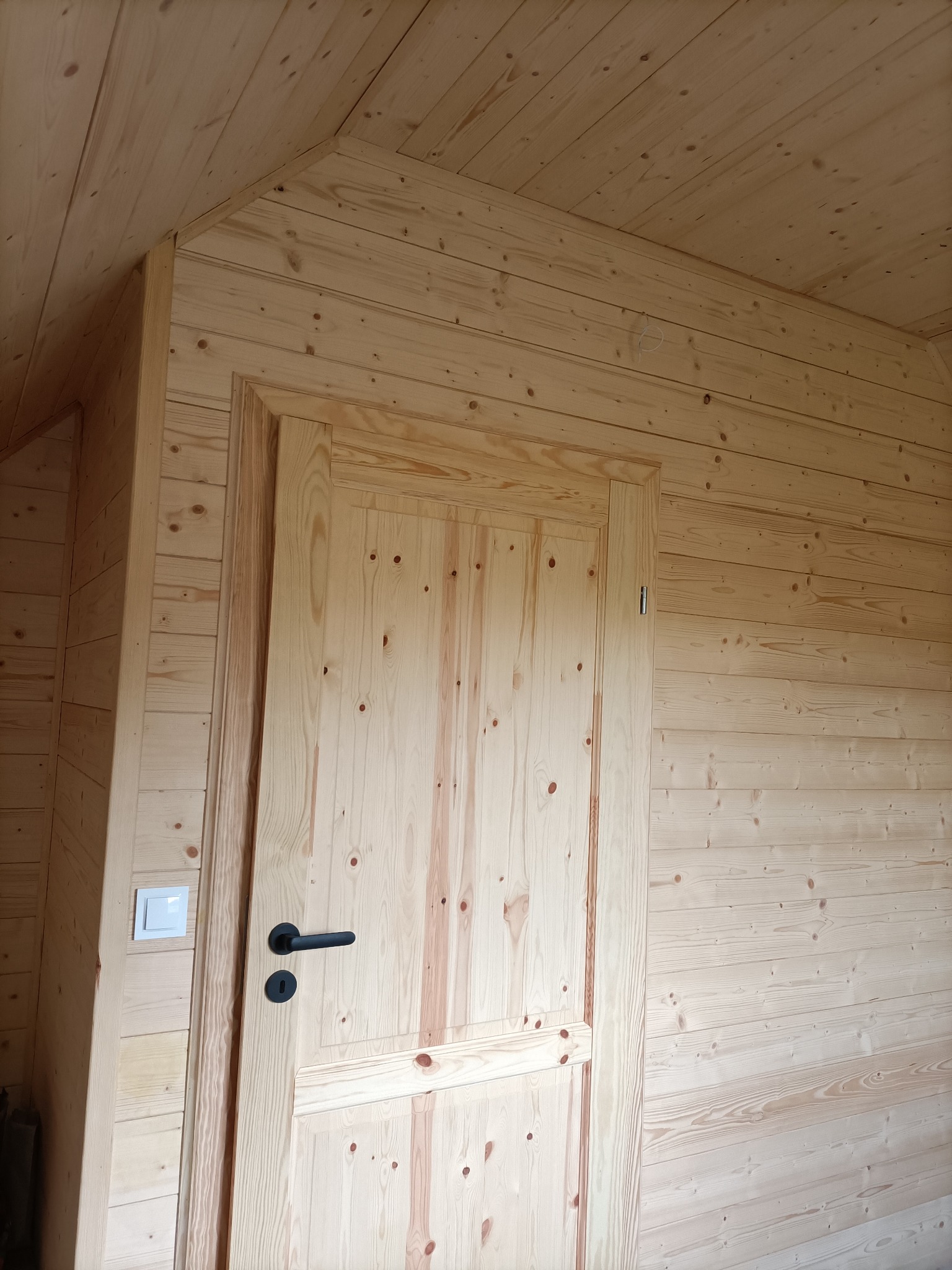 Wnętrze domu wykończone boazerią z jasnego drewna, widoczna drewniana rama drzwi z czarną klamką i włącznik światła po lewej stronie.