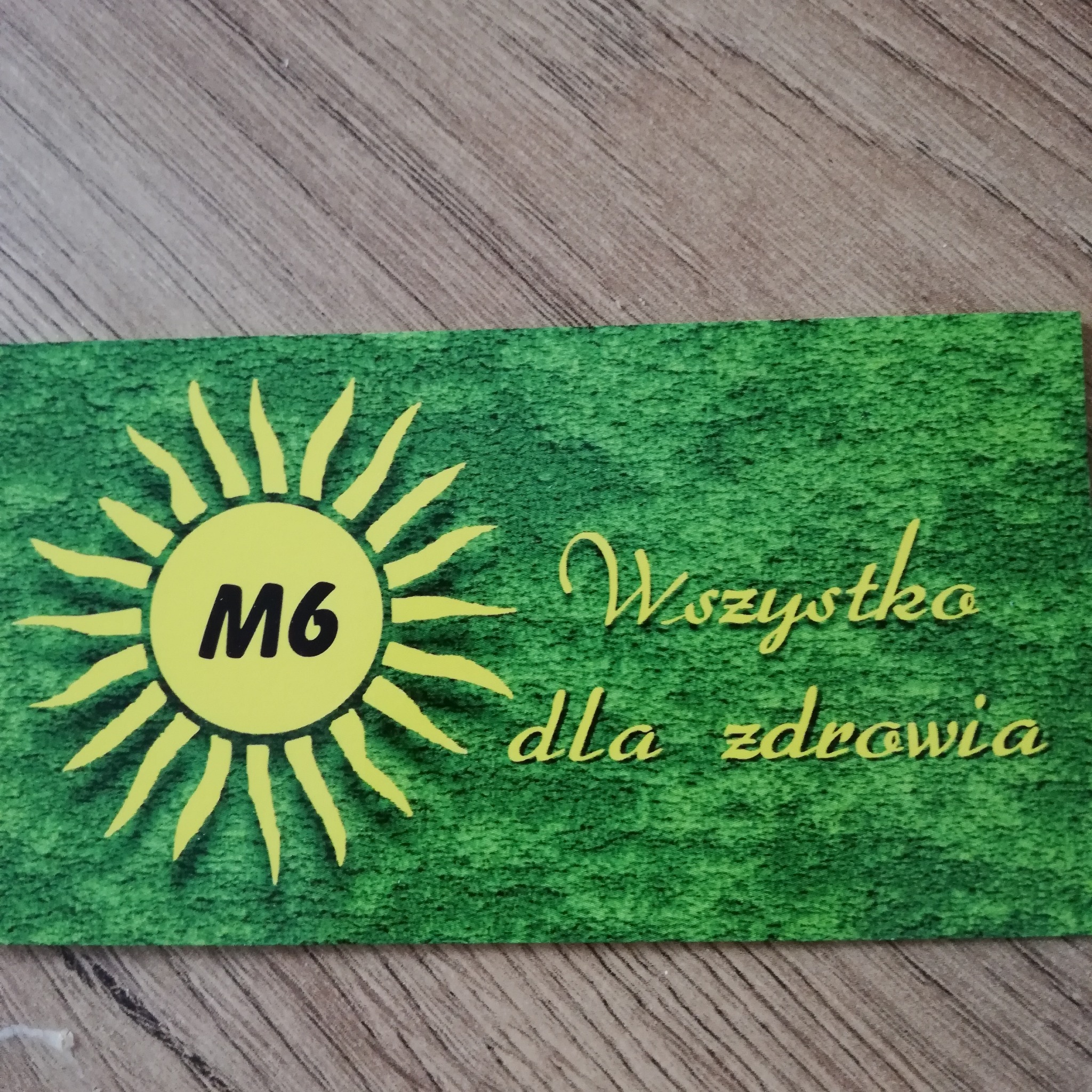 Karta z zielonym tłem i żółtym słońcem z napisem M6 oraz tekstem 'Wszystko dla zdrowia' leżąca na drewnianym stole.