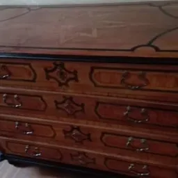 Komoda z ciemnego drewna z czterema szufladami, zdobiona czarnymi obramowaniami i metalowymi uchwytami, widoczna z perspektywy lekko z góry, stojąca na drewnianej podłodze w jasnym pomieszczeniu.