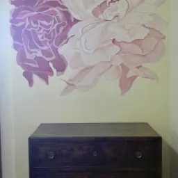 Ręcznie malowane, duże kwiaty piwonii w odcieniach różu i fioletu na kremowej ścianie nad ciemnobrązową, drewnianą komodą z dwoma szufladami w pomieszczeniu z parkietem.