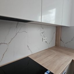 ELŻBIETA MIELNICKA ELIX - Kuchnia po remoncie: białe szafki wiszące, marmurowe płytki na ścianie, drewniany blat, czarna płyta indukcyjna i zlew.