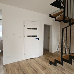 ELŻBIETA MIELNICKA ELIX - Jasne wnętrze z drewnopodobną podłogą, białą ścianą, nowoczesnymi schodami spiralnymi z metalową konstrukcją i drewnianymi stopniami oraz białe drzwi z poziomymi przeszkleniami, na ścianie widoczne...