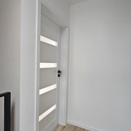 ELŻBIETA MIELNICKA ELIX - Nowoczesne, białe drzwi wewnętrzne z poziomymi przeszkleniami, czarna klamka, drewnopodobne panele podłogowe, białe ściany i sufit z czarnym reflektorem punktowym.