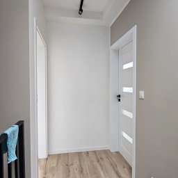 ELŻBIETA MIELNICKA ELIX - Korytarz po remoncie z drewnopodobną podłogą, białą ścianą, szarą ścianą z włącznikiem, białymi drzwiami z czarną klamką i otwartymi białymi drzwiami z widocznym fragmentem niebieskiej szmatki...