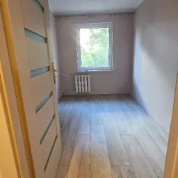 Jasne panele podłogowe imitujące drewno w pokoju z oknem i grzejnikiem. Widok zza otwartych drzwi z przeszkleniami.