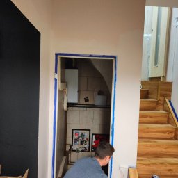 ELŻBIETA MIELNICKA ELIX - Mężczyzna maluje ścianę wałkiem w pomieszczeniu z drewnianymi schodami i wejściem do kotłowni, zabezpieczonym taśmą malarską.