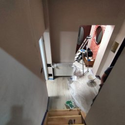 ELŻBIETA MIELNICKA ELIX - Klatka schodowa w trakcie malowania: ściany zabezpieczone taśmą malarską, widoczne schody drewniane, grzejnik i drabina malarska w tle, podłoga zabezpieczona folią malarską.