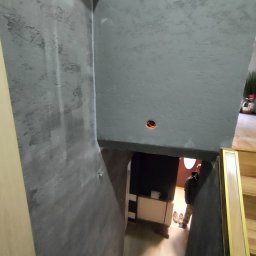 ELŻBIETA MIELNICKA ELIX - Szara ściana z fakturą w korytarzu ze schodami, widoczny otwór na oświetlenie w suficie i osoba w tle.