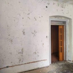 ELŻBIETA MIELNICKA ELIX - Pokój przed remontem: ściany z odpadającą farbą, widoczne ślady po poprzednich warstwach, drzwi wewnętrzne z drewnianą harmonijką, gniazdko elektryczne na ścianie, sufit z kasetonami.